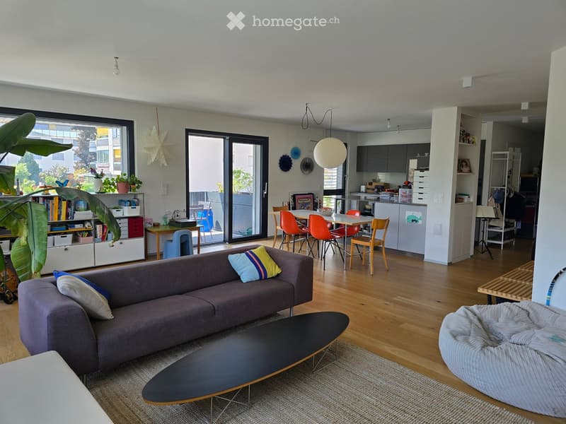A modern, open-concept living and dining area with a grey sofa, coffee table, beige chair, bookshelf, dining table with chairs, and a kitchen area with cabinets, counter, sink, stove, refrigerator, microwave, dishwasher, oven, and various kitchen items.