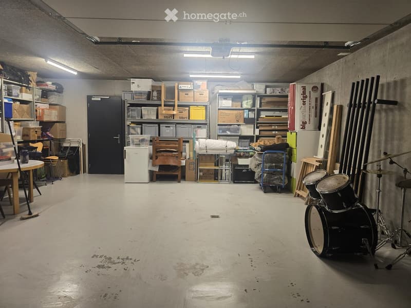 A spacious, well-lit storage or utility room with concrete floors and walls. Contains a drum set, shelves with storage boxes, a small refrigerator, a table with chairs, and various musical instruments and equipment.