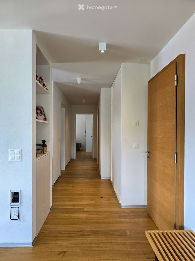 A long, well-lit hallway with wooden flooring, white walls, and recessed ceiling lights. Features built-in shelving on one side and a closed wooden door at the end.