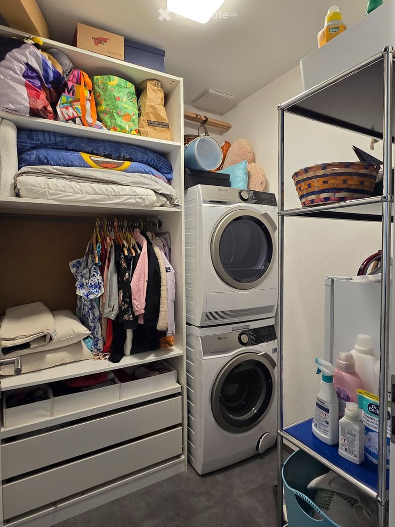 Well-organized laundry room with a washing machine, wardrobe with hanging clothes, shelves filled with bedding and bags, and cleaning supplies on a small cart.