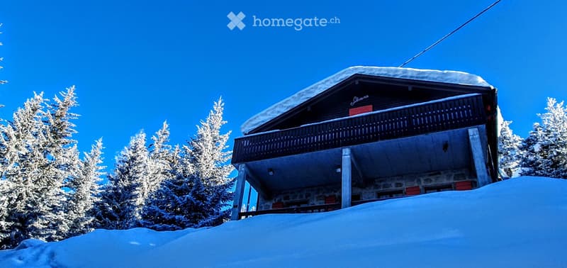 Two-story wooden chalet with a snow-covered roof and balcony, surrounded by snow-covered pine trees.