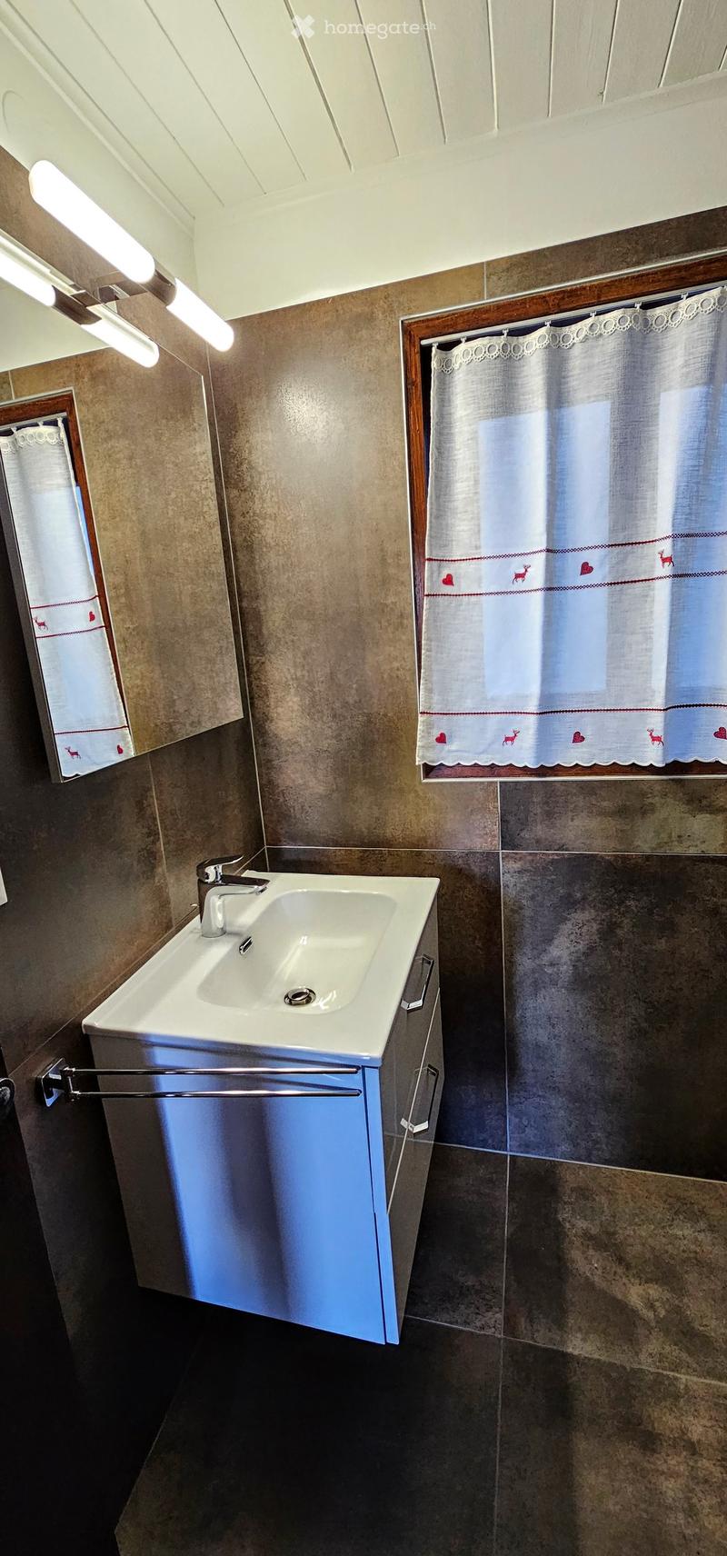 Bathroom with a white sink, chrome faucet, mirrored cabinet, and tiled walls. Light-colored curtains with deer and heart designs are visible on the windows.