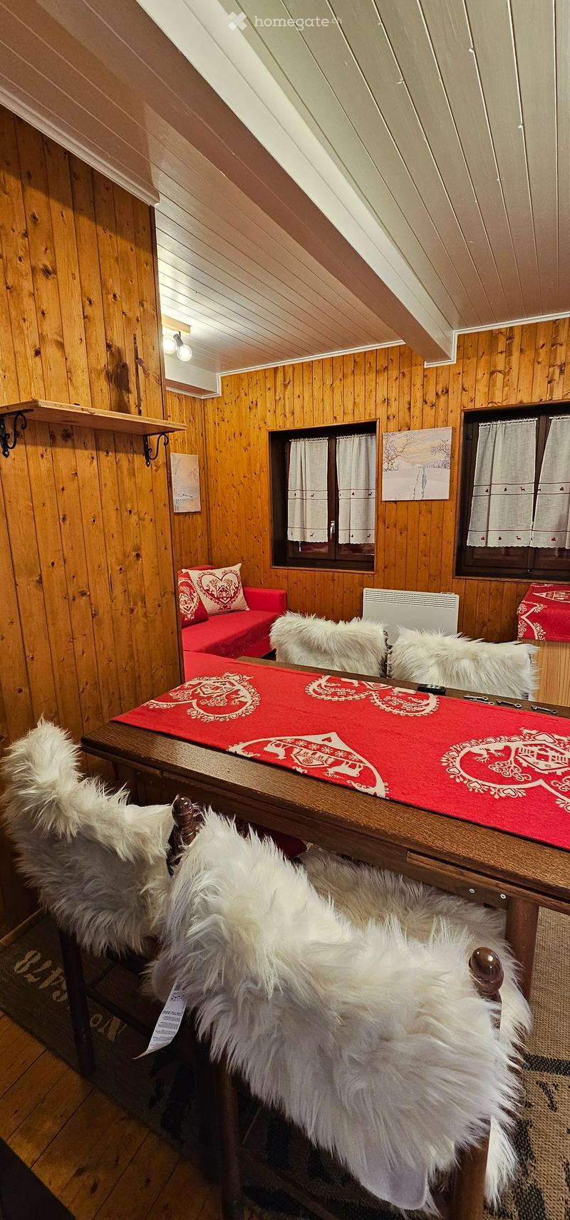 Cozy attic room with wooden walls and ceiling, featuring a red sofa, a wooden table with fur-covered chairs, and shelves with artwork.