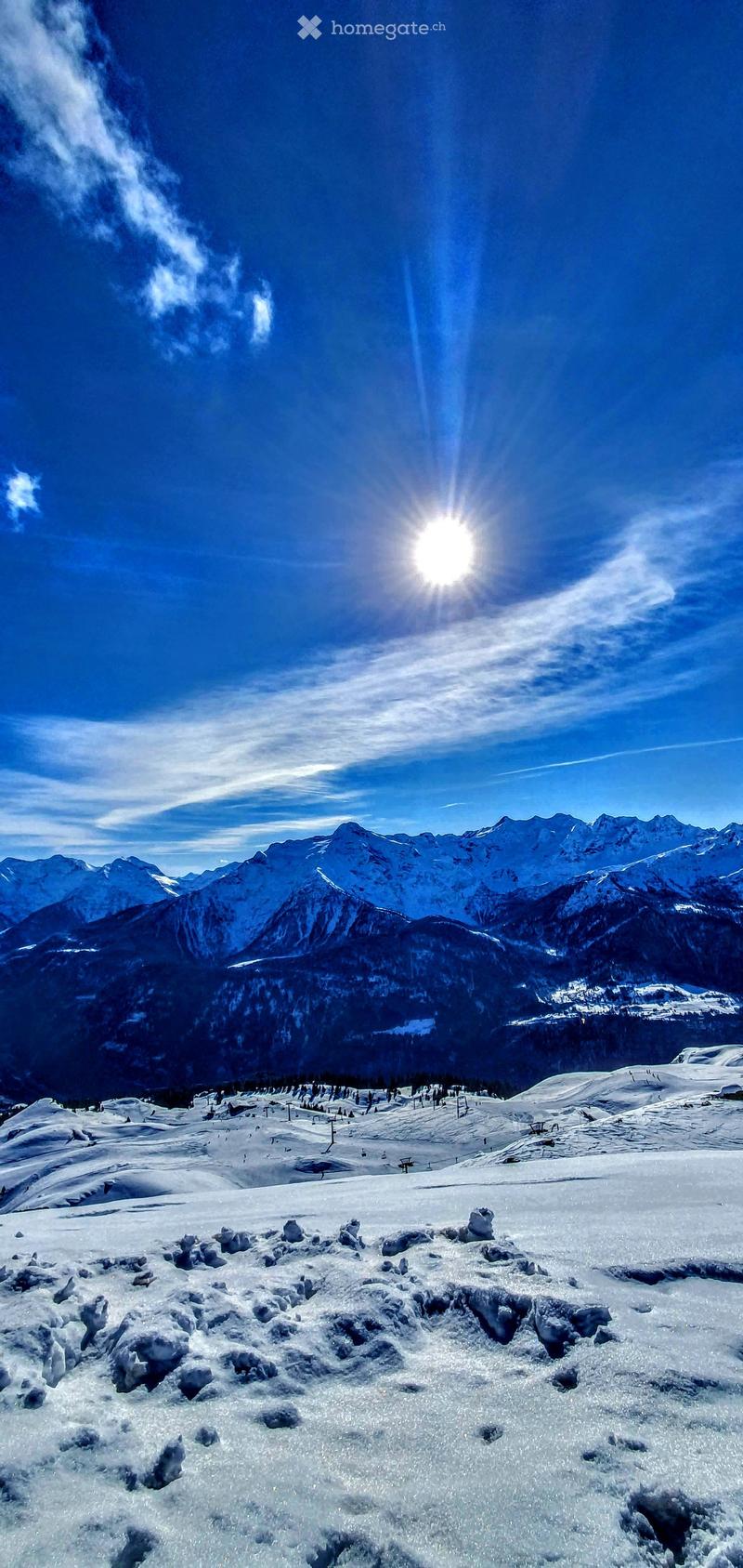 A snowy mountain landscape with a clear blue sky and sun shining brightly, featuring snow-covered peaks and a ski slope in the distance.