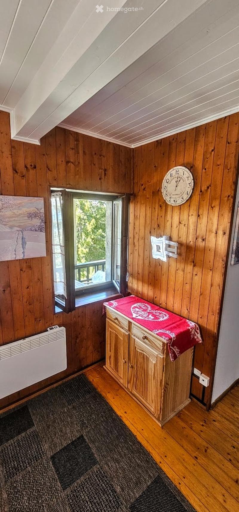 Cozy wooden-paneled room with a slanted ceiling, a window opening to a balcony, a small cabinet with a red cloth, a clock on the wall, and a radiator below the window.