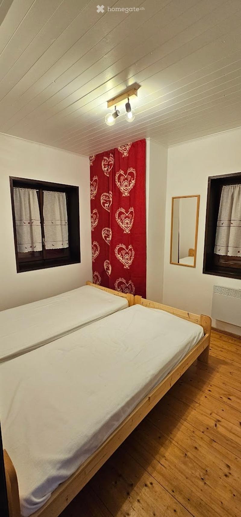 Cozy bedroom with two single beds, wooden flooring, white walls, and a red curtain with heart patterns. A small mirror is mounted on the wall, and the room is illuminated by ceiling lights.