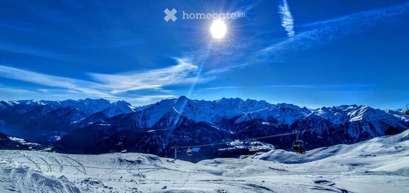 A panoramic view of snow-covered mountain peaks and a ski slope with a cable car system running through the landscape under a clear blue sky.