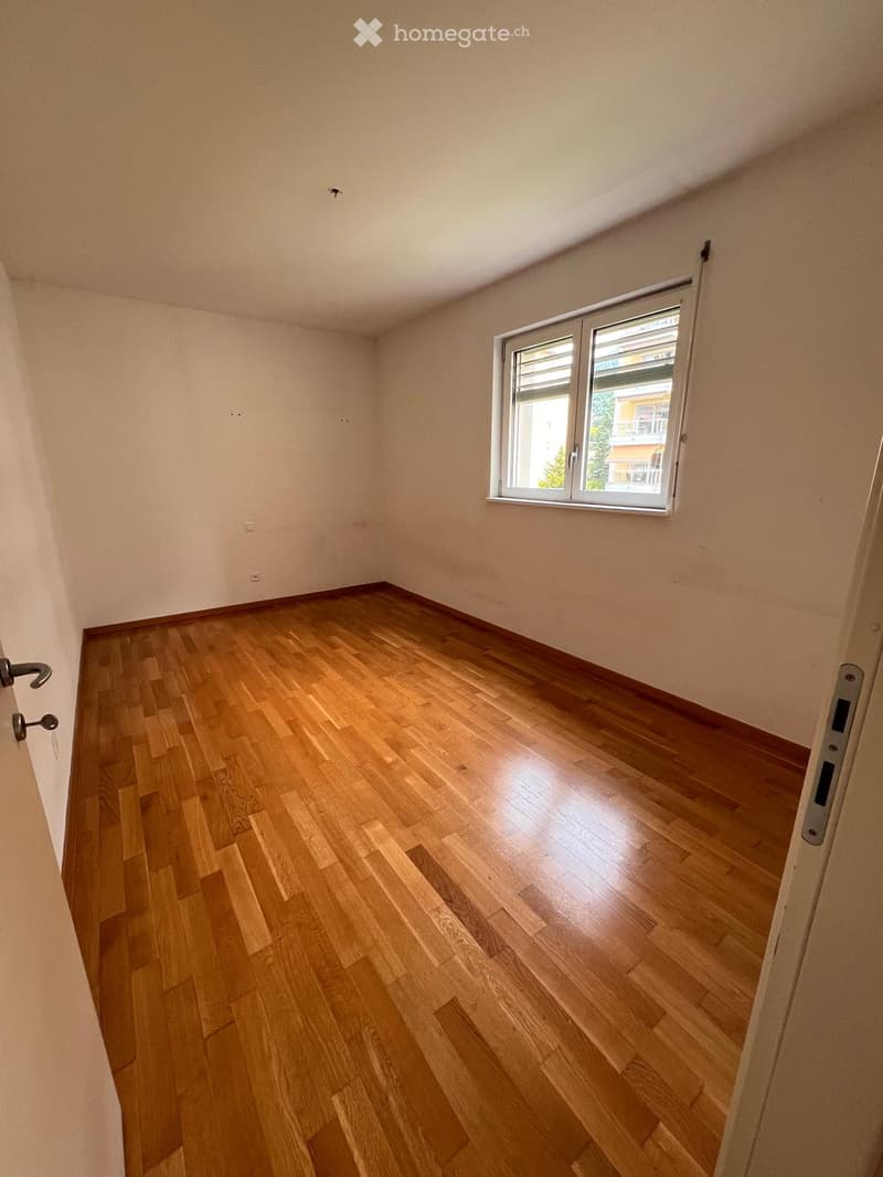 Empty room with wooden flooring and white walls, featuring a large window with blinds and a door with a handle visible on the left side.