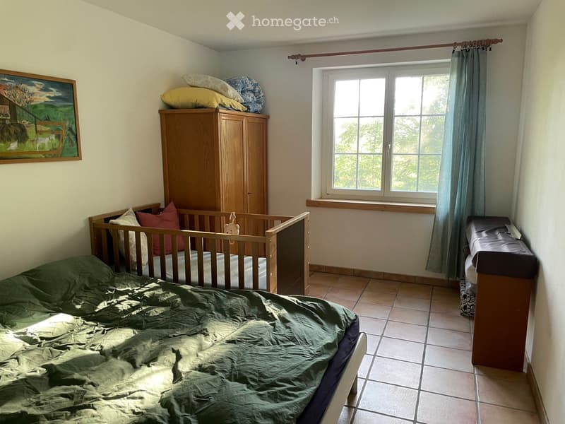 Cozy bedroom with a double bed, wooden wardrobe, small cradle, and a painting on the wall. The room has tiled flooring and a large window with curtains.
