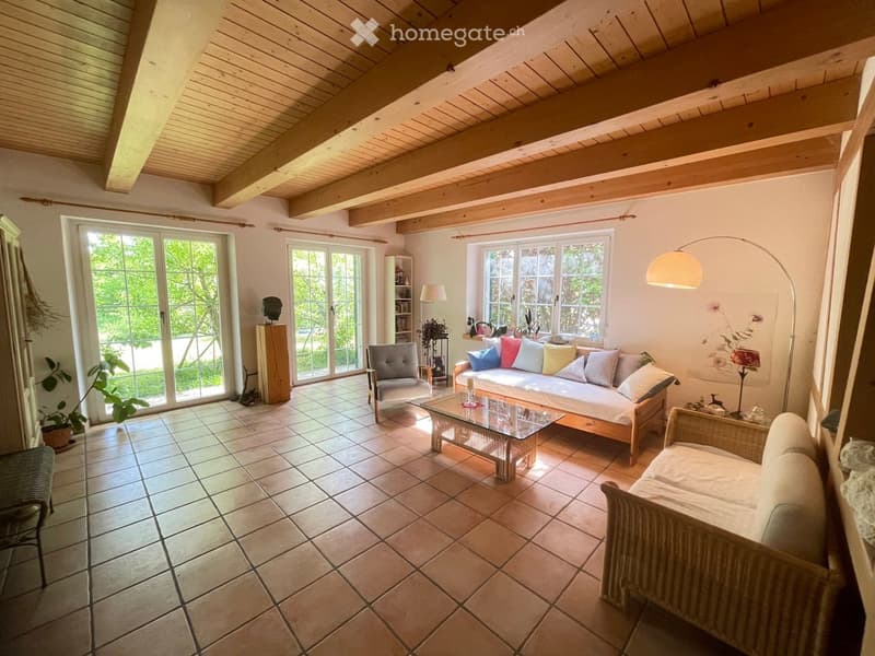 Bright living room with exposed wooden beams, large windows, and a mix of seating including a sofa, armchair, and wicker chair. The room features a glass coffee table, floor lamp, and a bookshelf with decorative items.