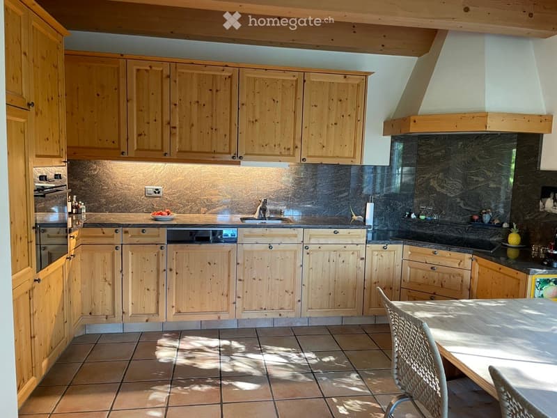 Bright kitchen with wooden cabinets, stainless steel appliances, and a central island with a sink and stove. The space features tiled flooring and a large window allowing natural light to fill the room.