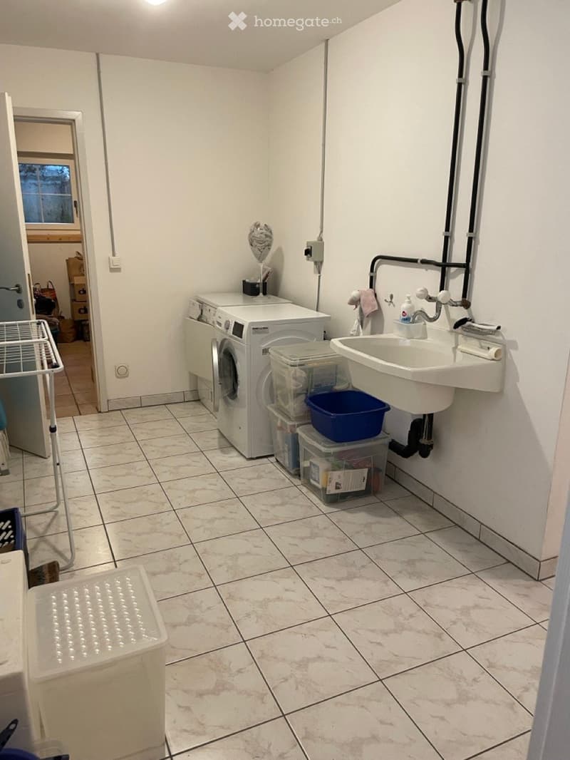 Laundry room with white tiled floor, washing machine, sink, plastic bins, and a mirror on the wall.