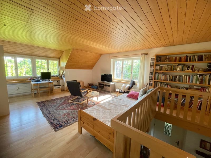 Bright living room with wooden beams, large windows, a sofa, chairs, a coffee table, a bookshelf, and a desk area.
