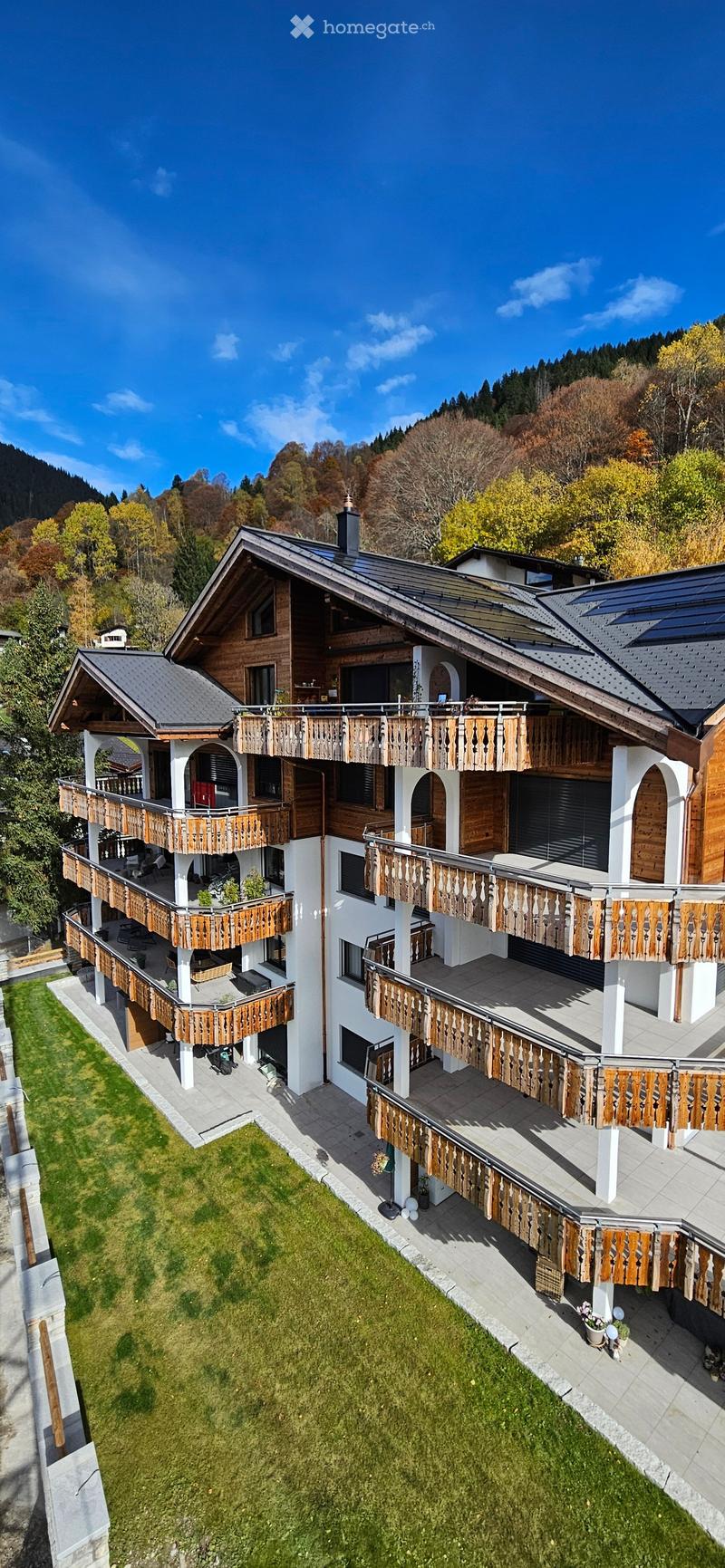 A large, multi-story wooden house with multiple balconies and a well-maintained lawn, surrounded by trees and mountains.