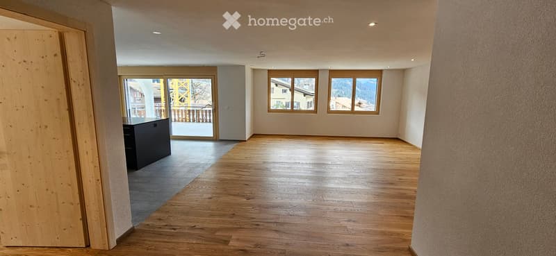 Empty room with wooden flooring, large windows, and a small black cabinet near the entrance. The space is well-lit with recessed ceiling lights.
