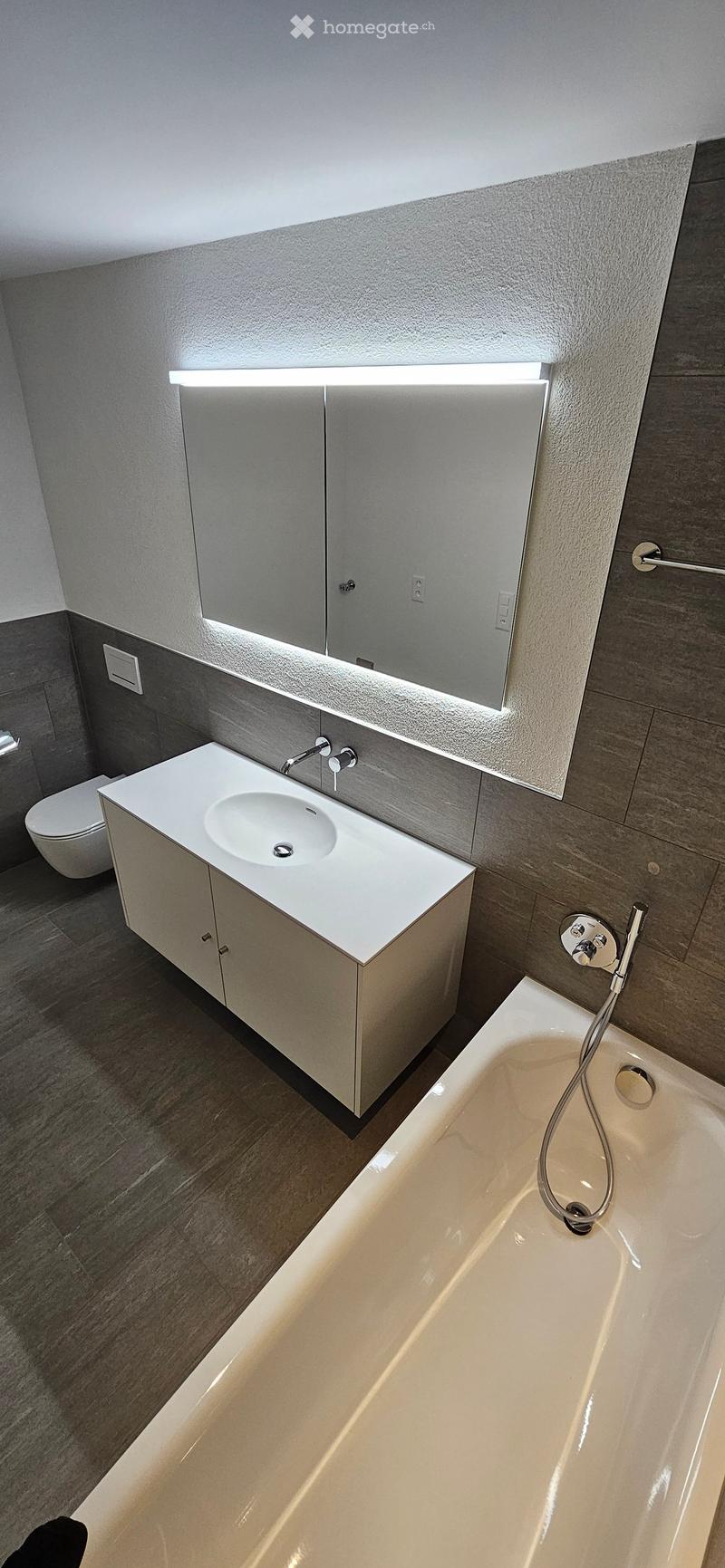 Modern bathroom with a white sink, bathtub, and large mirror with built-in lighting. The space features grey tiles and a sleek, contemporary design.