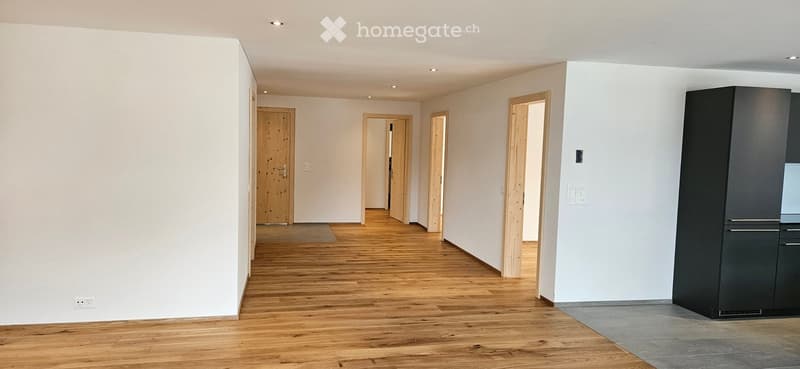 Empty hallway with wooden flooring, white walls, and several open doorways leading to other rooms. A small black cabinet is visible on the right side.