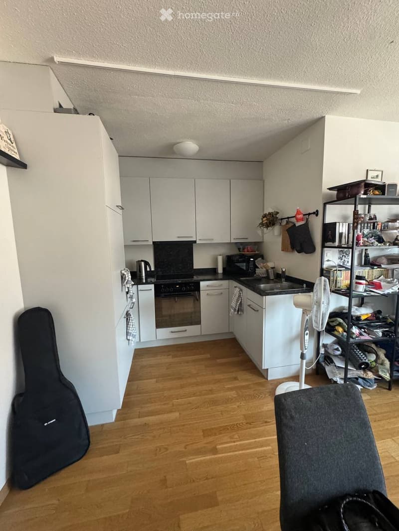 Modern kitchen with white cabinets, black countertops, and wooden flooring. Includes a refrigerator, oven, microwave, sink, fan, guitar case, and a shelf with various items.