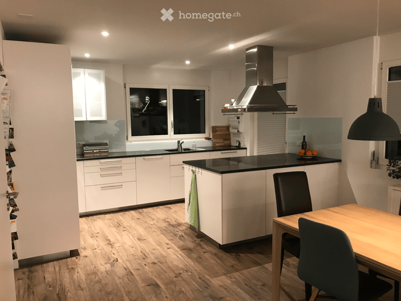 Modern kitchen with white cabinets, black countertops, wooden flooring, and a central island with a sink and range hood. Includes a dining area with a table and chairs.
