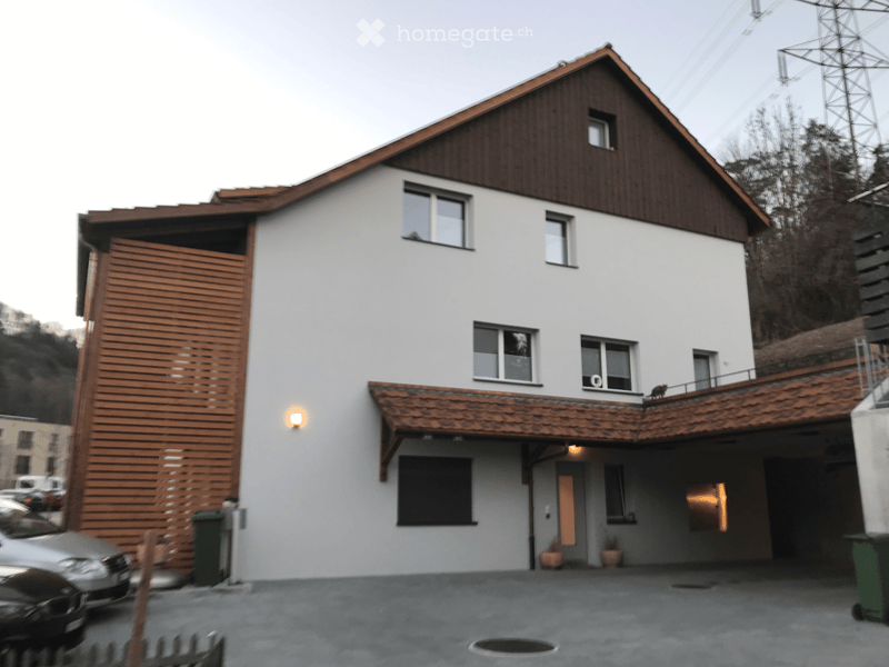 Two-story house with white walls, wooden roof, multiple windows, and a covered entrance area. Features a wooden garage door and a small balcony on the upper floor.