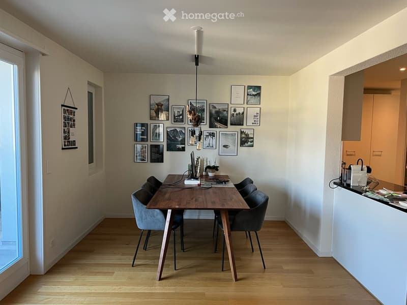 A modern, open-plan dining and kitchen area with a wooden dining table, six chairs, a screen, and a kitchen counter with cabinets. The walls are decorated with several picture frames.