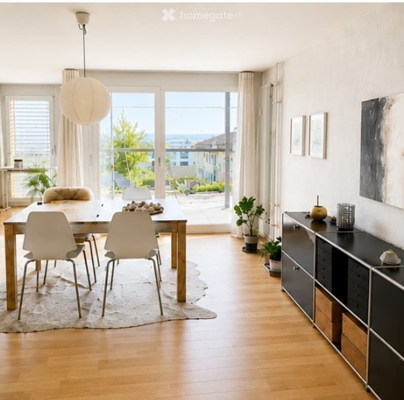 Bright dining area with wooden floor, large windows offering a sea view, modern dining table with four white chairs, sleek black cabinet, potted plants, and artwork on the walls.