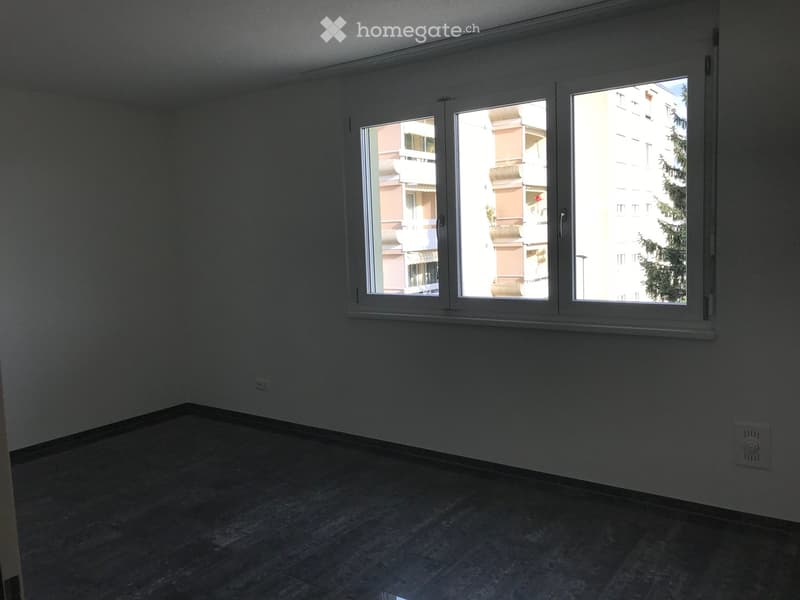Empty room with tiled floor, white walls, and large windows showing a view of neighboring buildings and greenery.