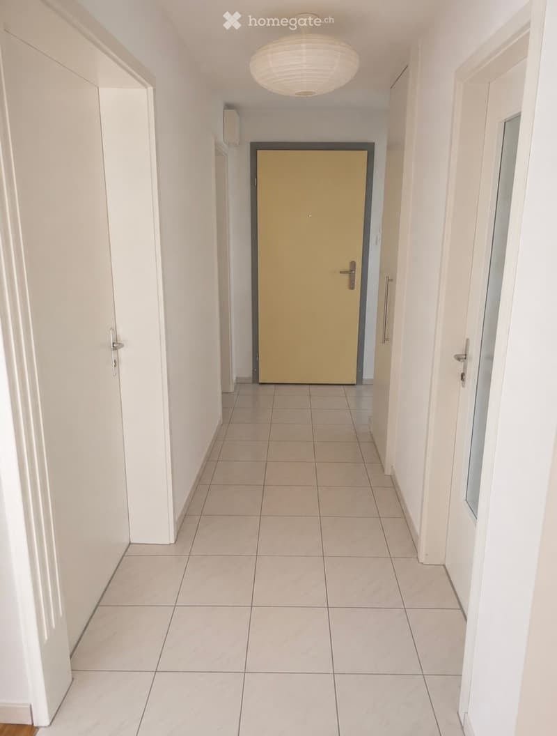 A clean, well-lit hallway with tiled flooring, white walls, and a central light fixture. A yellow door is visible at the end of the corridor.