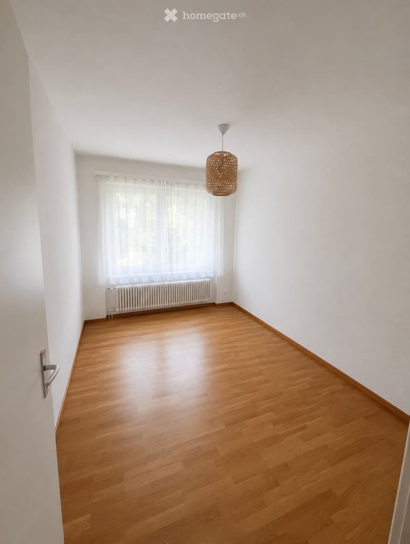 Empty room with wooden flooring, white walls, a window with white curtains, a radiator, and a hanging woven light fixture.