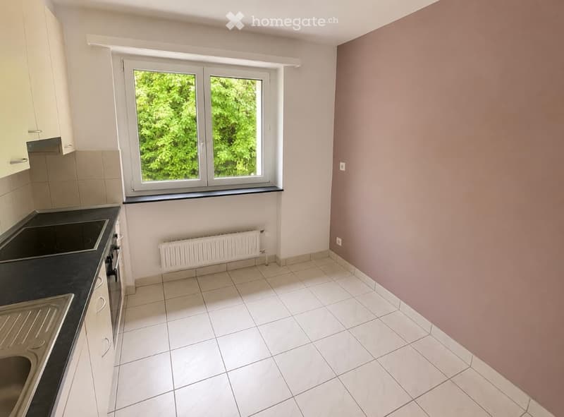 Empty kitchen with white tiled floor, white cabinets, black countertop, and a window overlooking greenery.