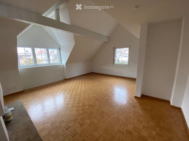 Empty attic room with wooden flooring, white walls, and large windows offering natural light. No furniture or equipment present.