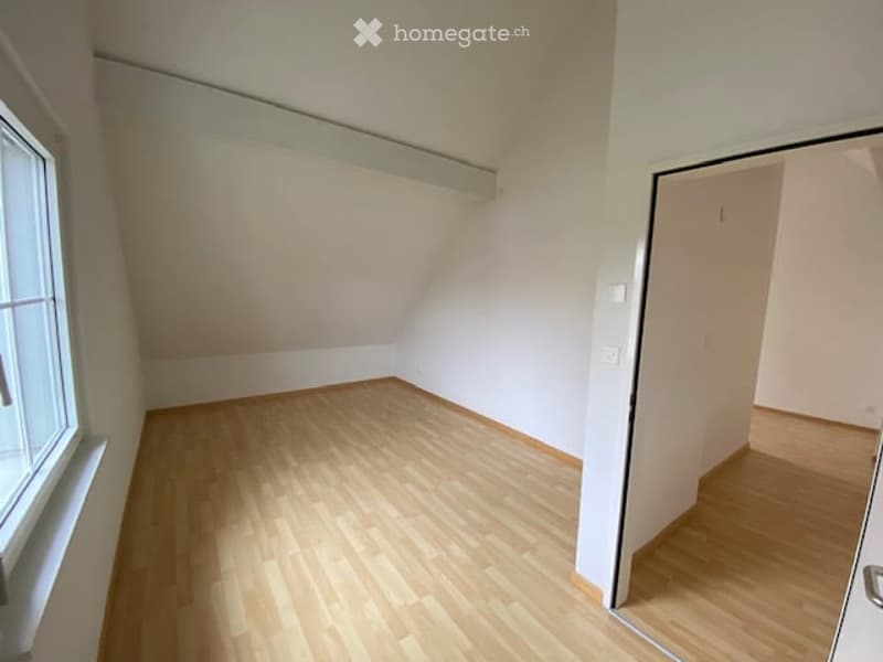 Empty room with wooden flooring, white walls, and a large window on one side. The room has a slanted ceiling typical of an attic space.