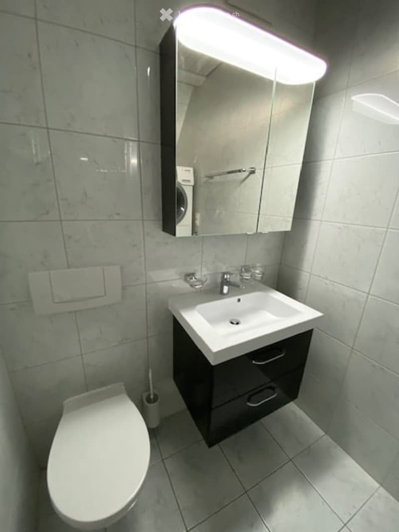 Modern bathroom with a white sink, toilet, and large mirror above the sink. The sink has a sleek black cabinet underneath, and the walls are tiled in a light marble pattern.