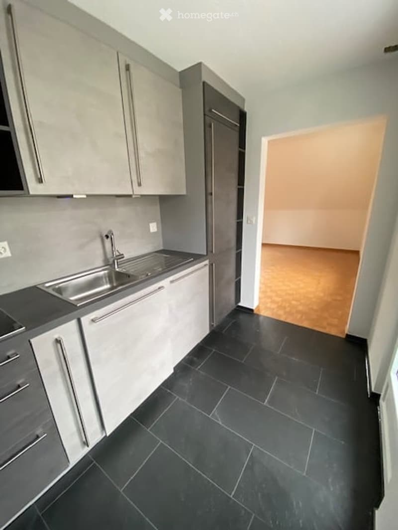 Modern kitchen with grey cabinets, black tiled floor, stainless steel sink, and a visible adjacent room with wooden flooring.