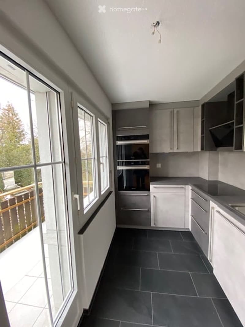 Modern kitchen with grey cabinets, black tiled floor, large windows, and built-in oven.