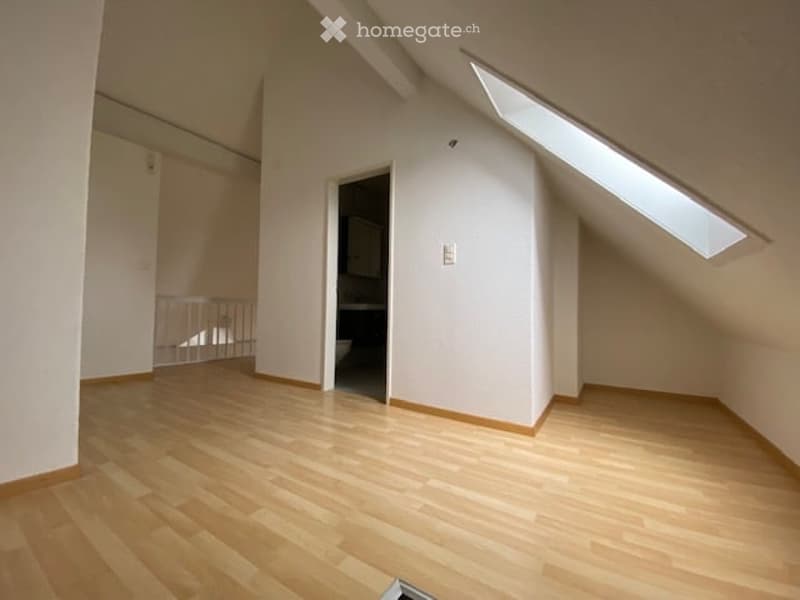 Empty room with wooden flooring, white walls, and a skylight window. A small bathroom is visible through an open doorway.