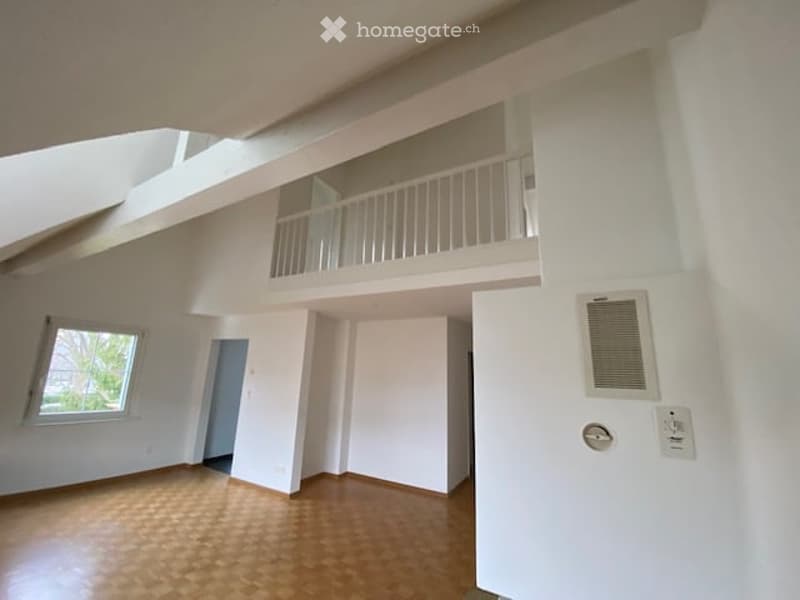Bright, open-plan room with wooden flooring, white walls, and a raised balcony area accessible through a doorway. The space features large windows and a modern, minimalist design.