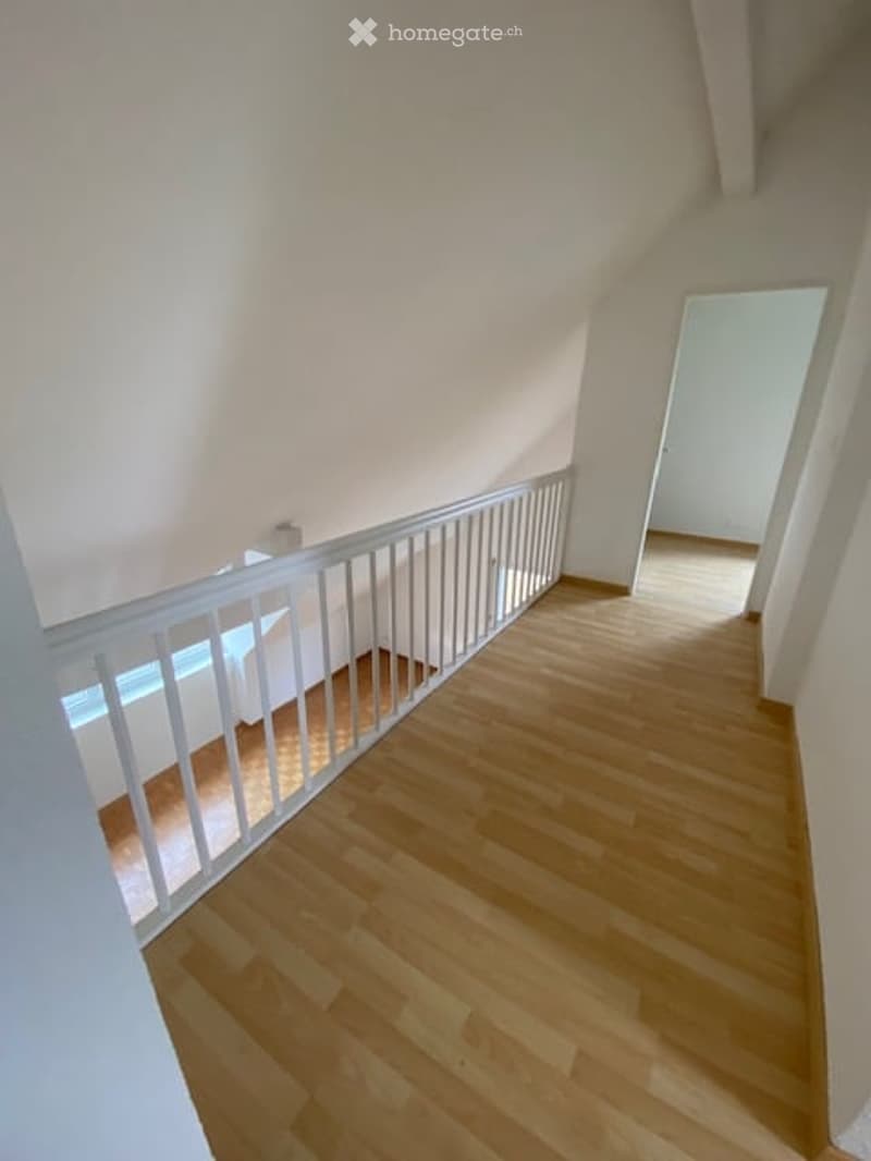 Empty hallway with wooden flooring and white walls, featuring a white metal railing and a doorway leading to another room.