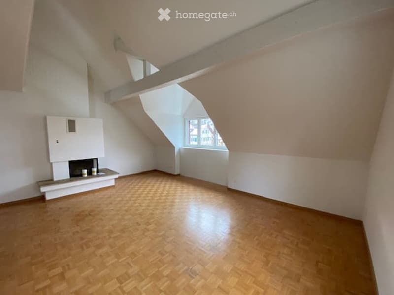 Empty attic room with wooden flooring, white walls, and a built-in fireplace. Slanted ceilings and a small window provide natural light.