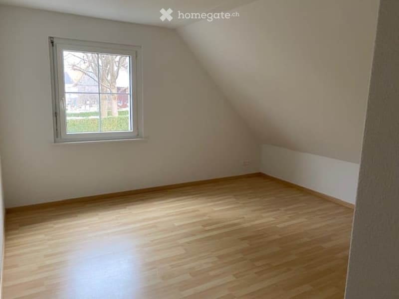Empty attic room with wooden flooring, white walls, and a single window showing a view of the outside.