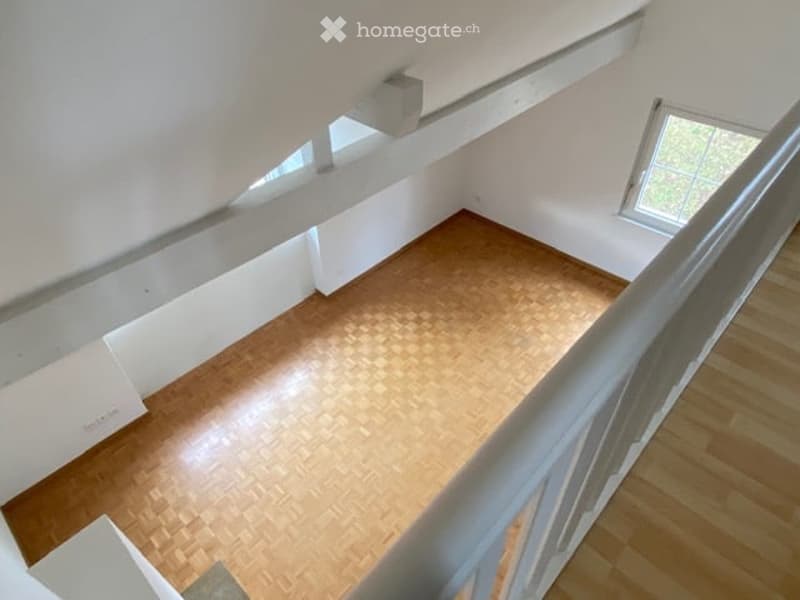 Empty room with wooden flooring and white walls, viewed from a staircase landing. No furniture or equipment present.