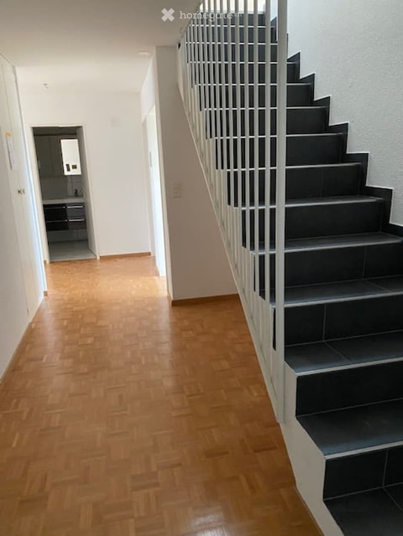 A modern hallway with wooden flooring, white walls, and a staircase with black steps and a metal railing. A small room with a sink is visible at the end of the hallway.