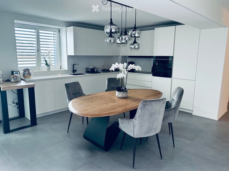 Modern kitchen with white cabinetry, a central wooden dining table with four grey chairs, pendant lights, and a sink area with a window and decorative flowers.