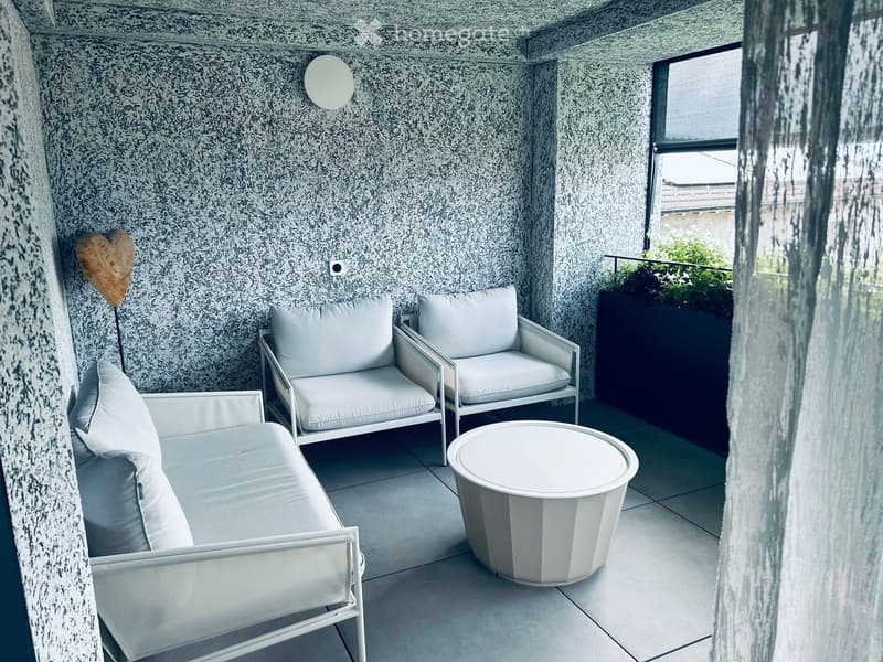 Modern balcony with white furniture including a sofa, two armchairs, and a round table. The walls are textured with a speckled pattern, and there is a large window with a view of greenery.