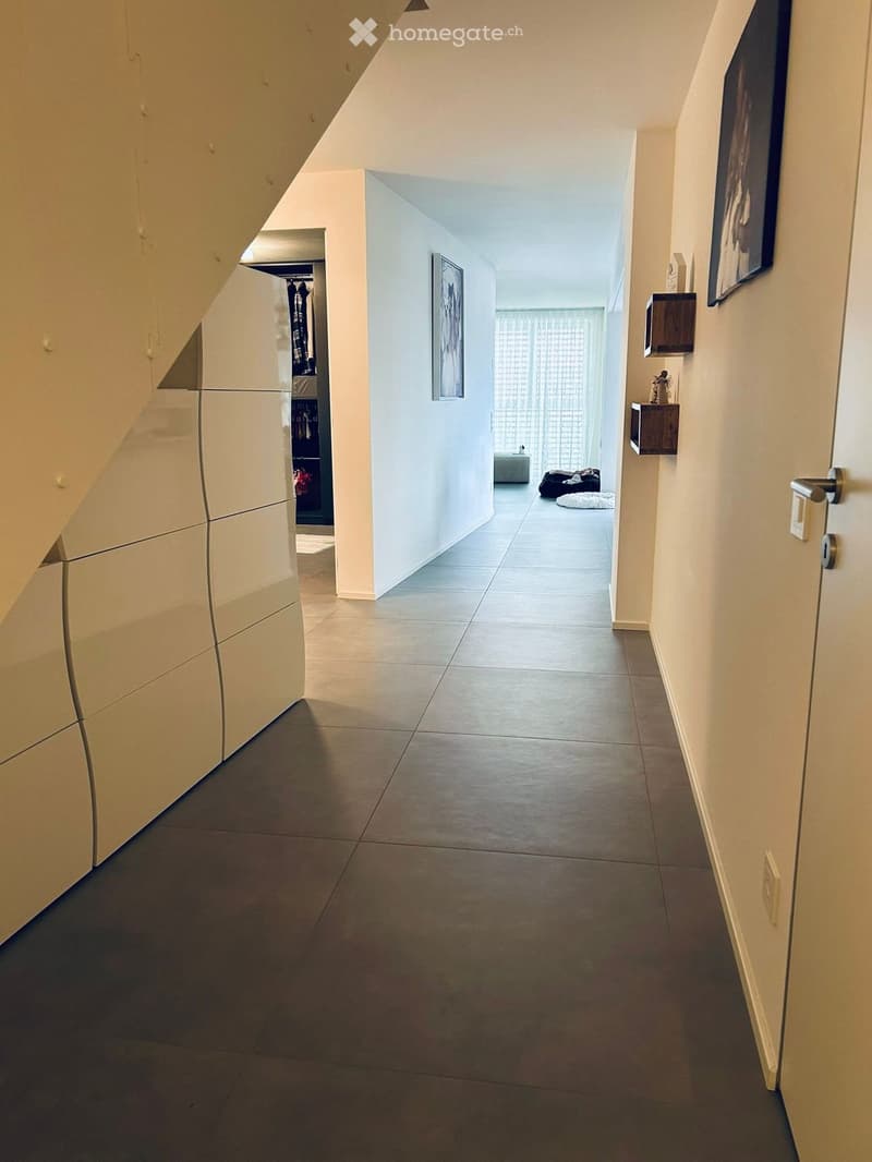Modern hallway with tiled floor, white walls, built-in closet on left, small shelf on right, and a glimpse of a room with a dog bed and a small table at the end.