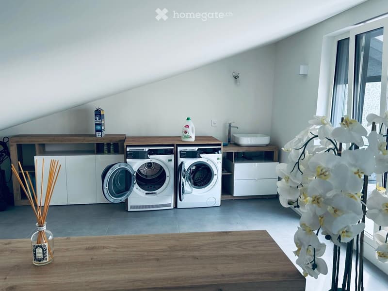 Modern laundry room with white washing machine and dryer, wooden cabinetry, sink, and decorative vase with white flowers.