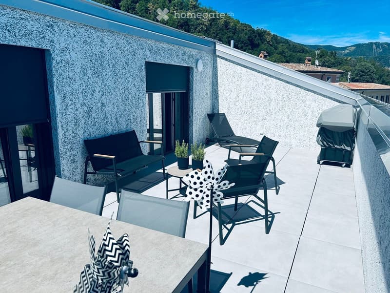 Modern rooftop terrace with a view of green hills, featuring a sofa, table, chairs, sun loungers, and an umbrella.
