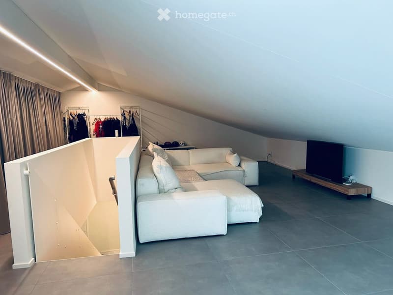 Modern living room with a white sectional sofa, a wooden TV stand, and a clothing rack with hanging clothes. The room features a sloped ceiling and large windows with curtains.