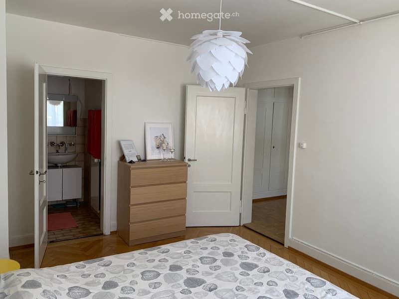 Bedroom with wooden floor, white walls, a bed with leaf-patterned bedding, a chest of drawers with picture frames and candles, an open door leading to a bathroom with a sink and mirror, and another open door leading to another room.