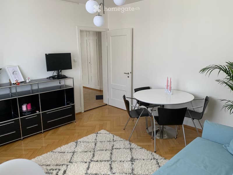 Modern living room with a light blue sofa, a white dining table with four chairs, a black TV cabinet, and a white shaggy rug on the wooden floor.